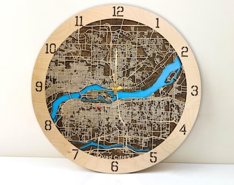 May include: A wooden clock with a map of the Quad Cities, Iowa, engraved on the face. The clock is 12 inches in diameter and has a light brown wood finish. The numbers are black and the hands are gold.