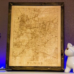 May include: A wooden framed map of Vilnius, Lithuania, with a detailed street map engraved on the wood. The map is in a brown frame and has the word "VILNIUS" printed in the bottom right corner.
