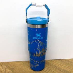 Puede incluir: Una taza de viaje de acero inoxidable azul con asa y tapa blanca. La taza tiene una escena de montaña y la palabra "STANLEY" impresa en ella.