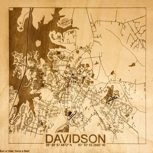 May include: A laser-engraved wooden map of Davidson, North Carolina. The map shows the town's streets, waterways, and landmarks. The text "DAVIDSON" is printed at the bottom of the map, along with the coordinates "35&deg; 29' 57.4872" N 80&deg; 50' 55.2696" W. The text "East or West, Home is Best!" is printed at the bottom left corner of the map.