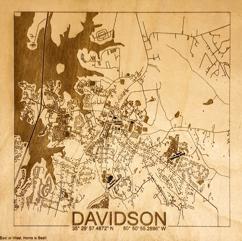May include: A laser-engraved wooden map of Davidson, North Carolina. The map shows the town's streets, waterways, and landmarks. The text "DAVIDSON" is printed at the bottom of the map, along with the coordinates "35&deg; 29' 57.4872" N 80&deg; 50' 55.2696" W. The text "East or West, Home is Best!" is printed at the bottom left corner of the map.