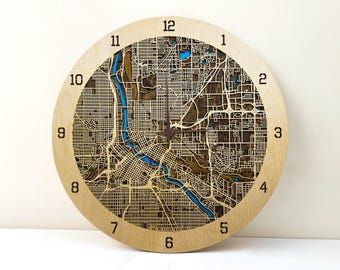 May include: A wooden clock with a laser-cut design of a city map. The clock face features the numbers 1 through 12 and a blue river winding through the city.