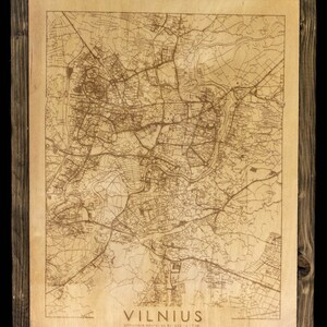 May include: A wooden framed map of Vilnius, Lithuania. The map is laser engraved on a light brown wood surface. The city name "VILNIUS" is printed in black at the bottom of the map.