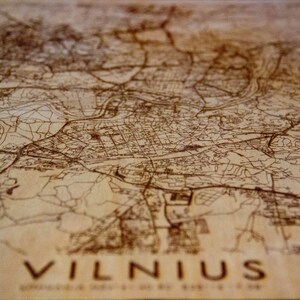 May include: A wooden map of Vilnius, Lithuania, with brown lines representing streets and waterways. The map is engraved with the word "VILNIUS" at the bottom.