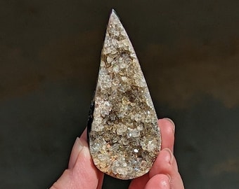 May include: A teardrop-shaped crystal geode with a sparkling, textured surface. The geode features a mix of clear, gold, and brown tones, with a dark, polished edge. The crystal is held in a hand against a blurred background.