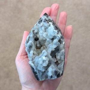 May include: A gray and white crystal point with a unique, textured surface. The crystal has a pointed top and a flat base.