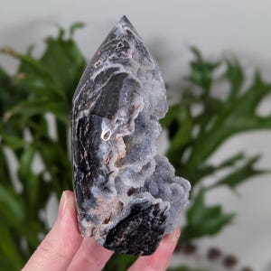 May include: Black and white agate crystal point with a natural druzy formation. The crystal has a pointed tip and a rough, textured surface.