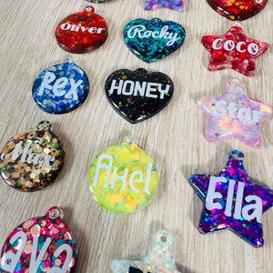 Op de afbeelding: Een verzameling kleurrijke dierenpenningen in diverse vormen, waaronder sterren, harten en cirkels. Elke penning is gevuld met glitter en voorzien van een dierennaam in witte letters. Namen: "Cooper", "Cardi", "Penny", "Oliver", "Rocky", "Coco", "Rex", "Honey", "Star", "Max", "Axel", "Ava", "Blue" en "Ella".