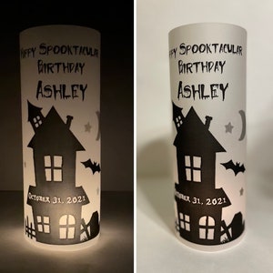May include: A white cylindrical candle with a black silhouette of a haunted house, bats, stars, and a crescent moon. The text "Happy Spooktacular Birthday Ashley" and "October 31, 2021" is printed on the candle.