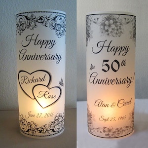 May include: Two white cylindrical candle holders with black and white designs. The first candle holder says "Happy Anniversary" with two hearts that say "Richard" and "Rose" and the date "June 27, 2016". The second candle holder says "Happy 50th Anniversary!" with the names "Alan & Carol" and the date "Sept 25, 1965".