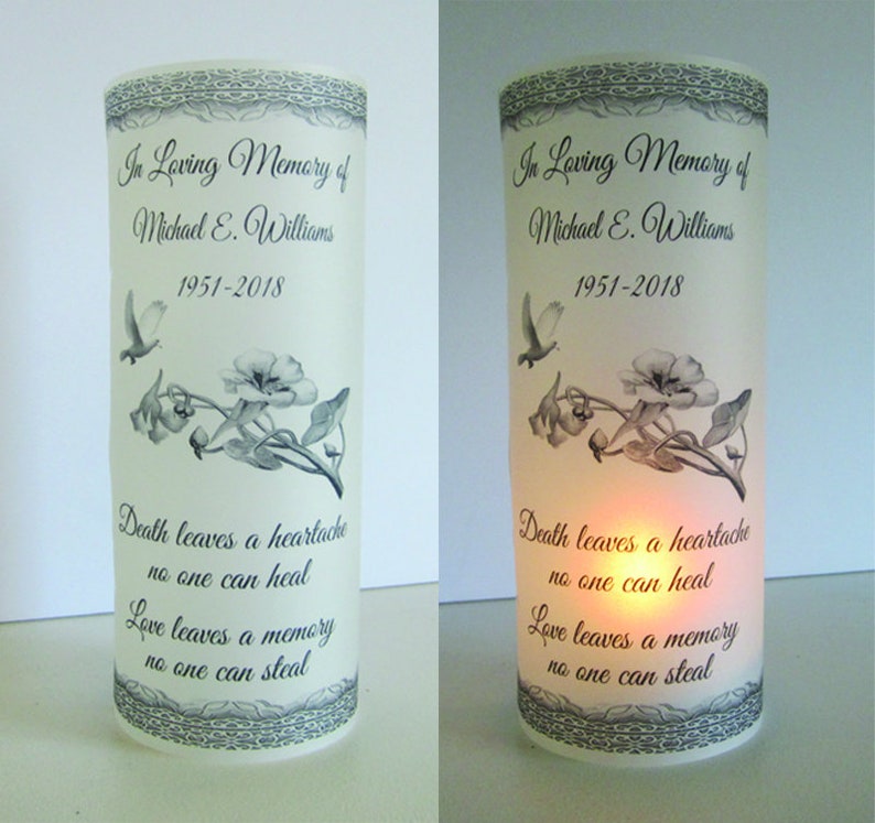 May include: A white pillar candle with a black and gray floral design and the text "In Loving Memory of Michael E. Williams 1951-2018" and "Death leaves a heartache no one can heal Love leaves a memory no one can steal."