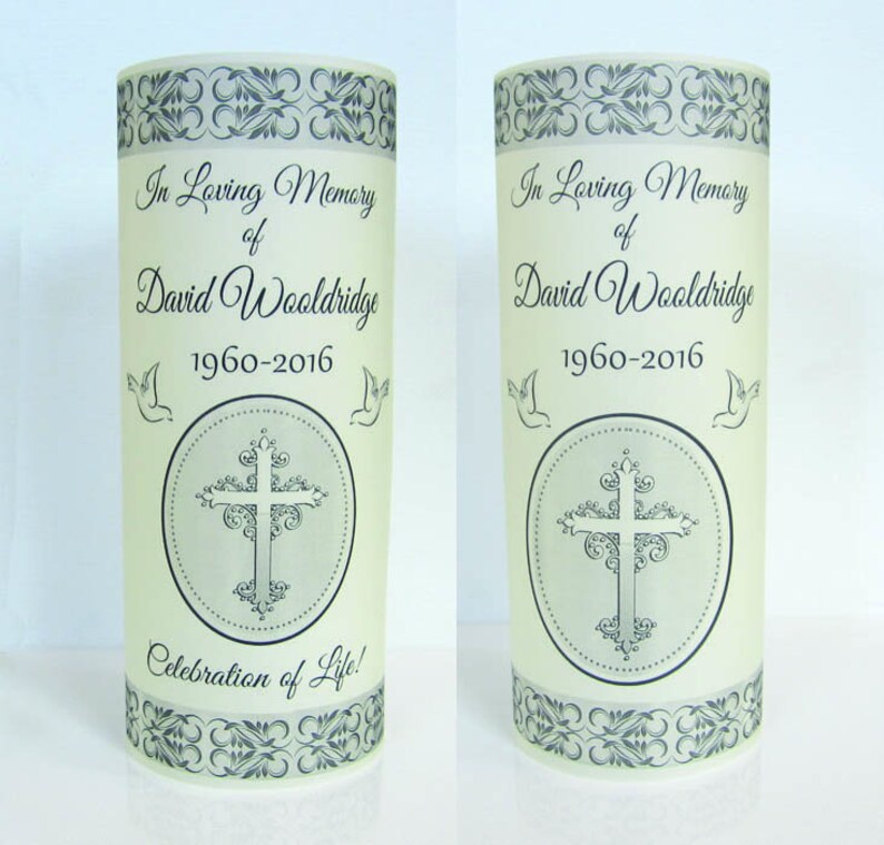 May include: Two white frosted glass candle holders with black decorative borders and a cross design. The text "In Loving Memory of David Wooldridge 1960-2016" and "Celebration of Life!" is printed on the candle holders.
