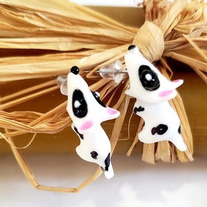 May include: A pair of white and black dalmatian dog stud earrings with pink accents. The earrings are made of polymer clay and have a realistic look.