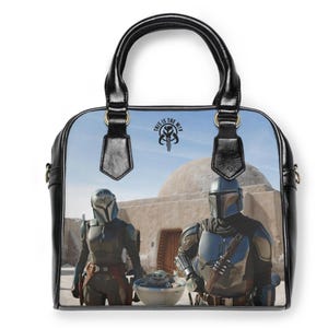 May include: Black handbag featuring a scene from a science fiction show. The bag has a black handle and trim, with a graphic of two characters in armor and a baby. The text "THIS IS THE WAY" is visible on the bag.