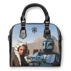 May include: A black handbag with a colorful Star Wars design. The bag features images of Ahsoka Tano, The Mandalorian, and Grogu. The text "THIS IS THE WAY" is printed on the bag. The bag has black handles and trim.