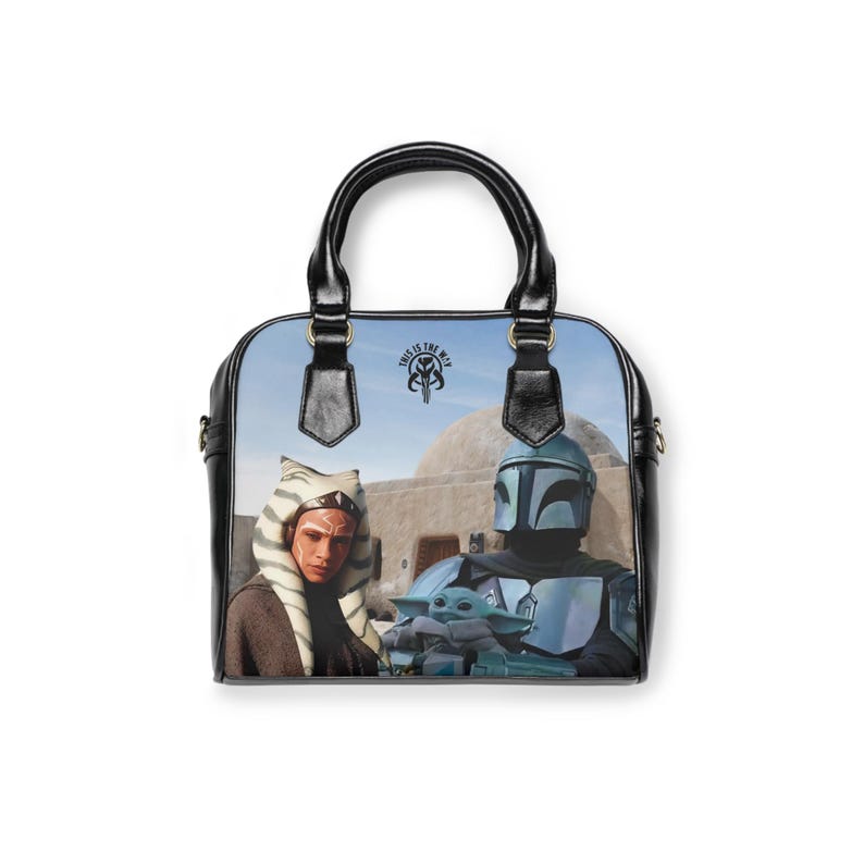 May include: A black handbag with a colorful Star Wars design. The bag features images of Ahsoka Tano, The Mandalorian, and Grogu. The text "THIS IS THE WAY" is printed on the bag. The bag has black handles and trim.