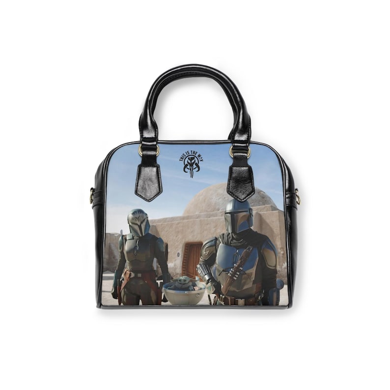 May include: Black handbag featuring a scene from a science fiction show. The bag has a black handle and trim, with a graphic of two characters in armor and a baby. The text "THIS IS THE WAY" is visible on the bag.