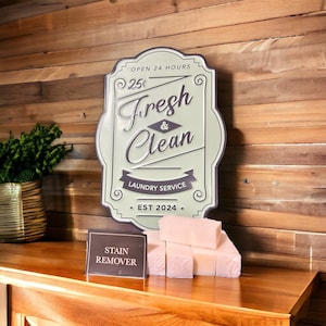 May include: A vintage-style metal sign with the text "Open 24 Hours 25¢ Fresh & Clean Laundry Service Est 2024." The sign is green with black lettering and is mounted on a wooden wall. There is a small sign that says "Stain Remover" and a stack of pink soap bars on a wooden surface.
