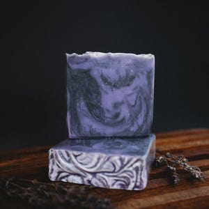 May include: Two bars of handmade soap with a purple and black marbled design. The soap is sitting on a wooden surface with dried lavender flowers.