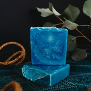 May include: Two bars of blue soap with a marbled design, sitting on a dark green textured surface.