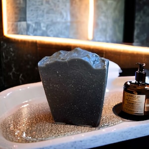 May include: A dark gray bar of soap in a white ceramic soap dish. The soap is wet and has a rough texture. A brown glass bottle with a pump dispenser is in the background.