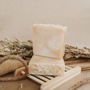 May include: Two bars of natural soap on a wooden soap dish. The soap is a light beige color with swirls of white. The soap dish is made of light brown wood and has a simple design.
