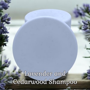 May include: Two round, light purple shampoo bars with the text "Lavender and Cedarwood Shampoo" in a soft, serif font. The bars are set against a blurred background with purple flowers.