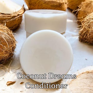 May include: Two white solid bars of Coconut Dreams Conditioner, a hair care product, are stacked on top of each other. The bars are on a white surface with coconut shells scattered around them.