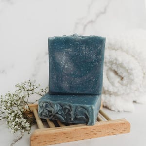 May include: Two bars of blue soap on a wooden soap dish. The soap is a dark blue color with a marbled pattern. The soap dish is made of light brown wood.