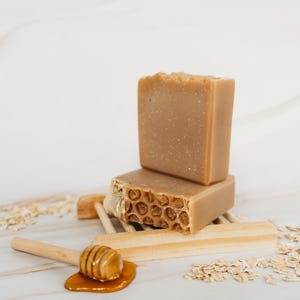 May include: Two bars of honey and oatmeal soap on a wooden soap dish with a honey dipper and oats scattered around.