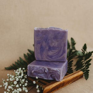 May include: Two bars of purple soap with swirls of white, sitting on a wooden soap dish. The soap is surrounded by small white flowers and green leaves.