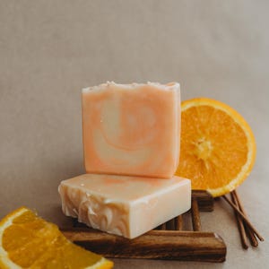 May include: Two bars of handmade soap with a swirl design, one pink and one white, on a wooden soap dish with orange slices and incense sticks.