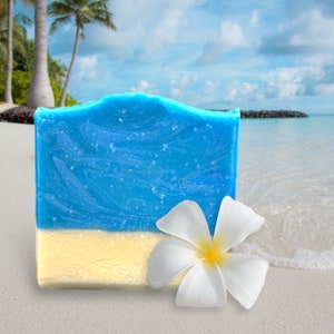 May include: A blue and white soap bar with a white plumeria flower on a sandy beach.