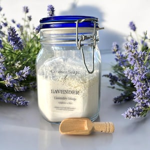 May include: A glass jar with a blue lid and a silver clasp filled with white laundry soap. The jar has a label that reads "Cerauno Soaps" and "Lavender Laundry Soap Approx. 2 lbs (Approx 17-18 loads HE washer / 37 loads regular washer)". A small wooden scoop is in front of the jar.