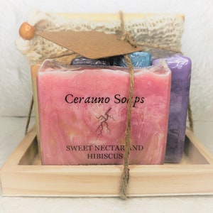 May include: A set of four handmade soap bars in a wooden tray. The soap bars are wrapped in a natural twine and tied with a brown paper tag that reads "Cerauno Soaps". The soap bars are pink, purple, and blue. The pink soap bar has the text "Sweet Nectar and Hibiscus" printed on it.