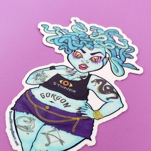 May include: A sticker featuring a cartoon illustration of a blue-skinned woman with snakes for hair. She is wearing a black tank top with the word "STONER" on it, a purple skirt, and high-heeled shoes. The text "GORGON" is written on her skirt. The text "Still Stunning" is written on her leg.