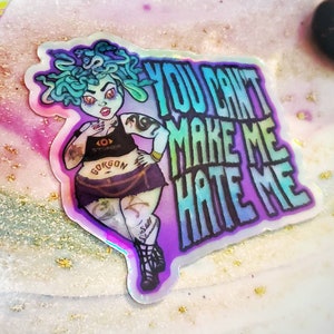 May include: A holographic sticker featuring a cartoon illustration of a green-haired woman with a snake-like head. She is wearing a black top with the word "Gorgon" on it and a purple skirt. The sticker has the text "You can't make me hate me" in a rainbow gradient.