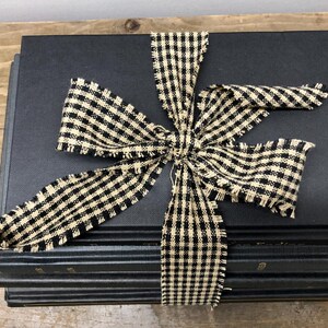 Pu&ograve; includere: Una pila di libri neri legati con un fiocco di nastro a scacchi crema e nero. Il fiocco ha bordi sfilacciati, che aggiungono un tocco rustico. I libri sono impilati su una superficie di legno.