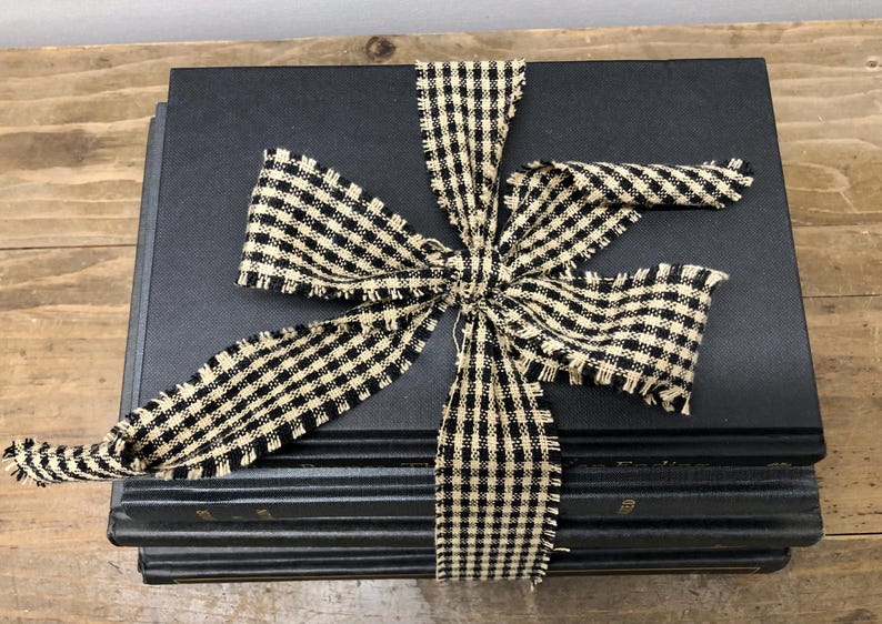 Pu&ograve; includere: Una pila di libri neri legati con un fiocco di nastro a scacchi crema e nero. Il fiocco ha bordi sfilacciati, che aggiungono un tocco rustico. I libri sono impilati su una superficie di legno.