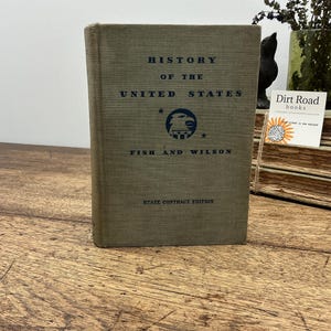 May include: A hardcover book titled History of the United States by Fish and Wilson, State Contract Edition. The cover is a muted olive-gray fabric with dark blue text and a central circular emblem featuring an eagle and stars.