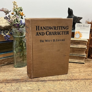 May include: A tan hardcover book titled Handwriting and Character by De Witt B. Lucas. The book features a textured fabric cover with black text. It is positioned upright among other vintage books, a glass vase with dried flowers, and a small sign.