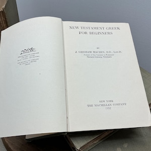 May include: Open book titled "New Testament Greek for Beginners" by J. Gresham Machen, published in 1952 by The Macmillan Company, New York. The book is open to the title page, with text in black on a cream-colored page. The Macmillan Company logo is also visible.