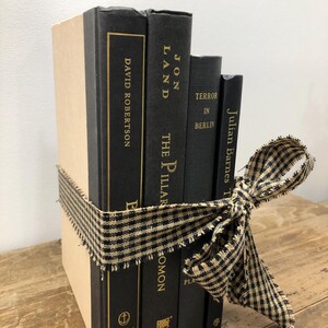 Pu&ograve; includere: Un gruppo di libri decorativi legati con un nastro a quadretti neri e beige. I libri hanno copertine nere con scritte dorate e titoli come "David Robertson" e "Jon Land". I libri sono disposti su una superficie di legno.