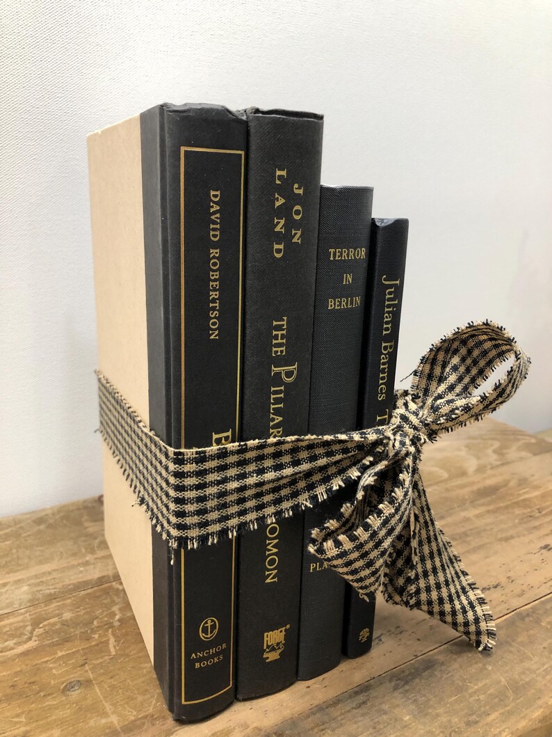 Pu&ograve; includere: Un gruppo di libri decorativi legati con un nastro a quadretti neri e beige. I libri hanno copertine nere con scritte dorate e titoli come "David Robertson" e "Jon Land". I libri sono disposti su una superficie di legno.