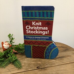 May include: A book titled "Knit Christmas Stockings!" in the shape of a Christmas stocking. The cover features a knitted pattern in shades of blue, green, and red. The book is on a wooden surface, with a small Santa figurine and greenery in the background.
