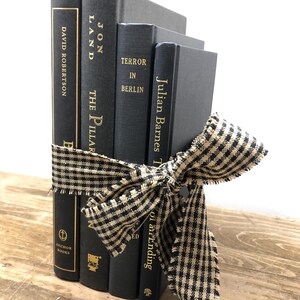 Pu&ograve; includere: Un gruppo di quattro libri neri legati con un nastro a quadretti neri e beige. I titoli dei libri includono "David Robertson", "Jon Land The Pillar", "Terror in Berlin" e "Julian Barnes T". I libri sono su una superficie di legno.