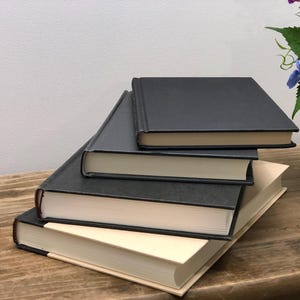 Pu&ograve; includere: Una pila di quattro libri con copertina rigida con copertine grigio scuro e pagine color crema. I libri sono disposti su una superficie di legno, con un vaso di fiori sullo sfondo. I libri sono di dimensioni variabili.