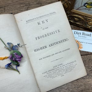 May include: An open antique book titled Robinson's Mathematical Series, Key to the Progressive Higher Arithmetic, for Teachers and Private Learners, published in 1862 in New York. The cream-colored page features black serif text and pressed flowers.