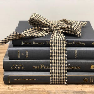 Pu&ograve; includere: Una pila di quattro libri grigio scuro legati con un nastro a scacchi beige e nero. I libri hanno scritte dorate e titoli come "The Fan Ending" e "Terror in Berlin". I libri sono impilati su una superficie di legno.