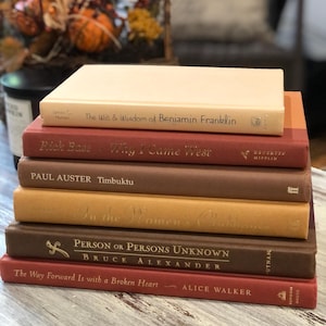 May include: A stack of seven hardcover books in various shades of brown, red, and beige. Titles include "The Wit & Wisdom of Benjamin Franklin," "Why I Came West," and "Timbuktu." The books are arranged on a light-colored wooden surface.